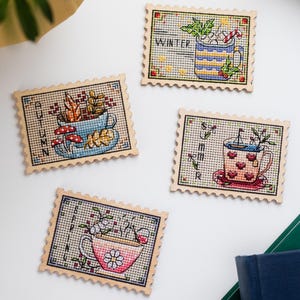 May include: Four cross-stitch coasters with seasonal designs. Each coaster is in a postage stamp shape and features a teacup with details representing winter, autumn, summer, and spring. The coasters are on a white surface with a plant and books in the background.