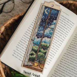 Könnte beinhalten: Handgefertigtes Lesezeichen aus Holz mit einem Kreuzstichmuster, das eine Nachtszene durch ein Fenster mit Mond, Sternen und Landschaft darstellt. Das Lesezeichen liegt auf einem offenen Buch. Der Text "Sweet Annet" ist sichtbar.