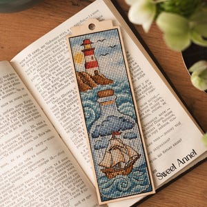 Könnte beinhalten: Bestickter Holz-Lesezeichen mit einem Leuchtturm, einem Schiff in einer Flasche und einem Segelboot auf blau-weißem Hintergrund. Das Lesezeichen liegt auf einem offenen Buch.