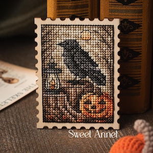 Peut inclure: Une pièce de point de croix rectangulaire à bord festonné représentant un corbeau noir perché sur un poteau en bois à côté d'une lanterne allumée et d'une citrouille sculptée orange. Le motif inclut une petite lune et le texte Sweet Annet.