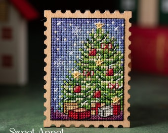 May include: A rectangular wooden frame with scalloped edges containing a cross-stitch embroidery of a Christmas tree decorated with red ornaments and yellow stars against a blue snowy background, with the text Sweet Annet printed at the bottom.