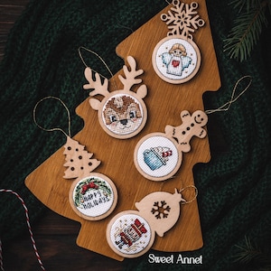 May include: Wooden Christmas ornaments with cross-stitch designs, including a reindeer, angel, gingerbread man, and nutcracker. The ornaments are arranged on a wooden tree-shaped board. The text "Happy Holidays" is visible on one ornament. The image is set against a dark green background.