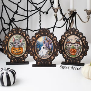 May include: Three framed Halloween cross-stitch artworks. The artworks feature a bat in a pumpkin, a ghost in front of a castle, and two skeletons with a pumpkin. The frames are dark brown with decorative edges. A black and white pumpkin and a white pumpkin are also present.