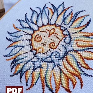 May include: A cross-stitch embroidery of a stylised sun with a face, surrounded by blue, yellow and orange rays. The embroidery is on a light blue fabric and is framed in a wooden hoop.