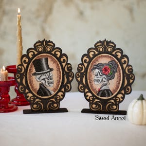 May include: Two framed cross-stitch portraits of skeletons, one in a top hat and suit, the other with a rose in her hair. The frames are dark brown with ornate designs. The background is a neutral color, with candles and a small pumpkin.