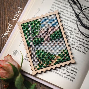 Könnte beinhalten: Ein rechteckiges Kreuzstich-Ornament aus Holz in Form einer Briefmarke mit einer Landschaftsdarstellung, die einen hohen Baum, grünes Blattwerk und Berge zeigt. Der Text Sweet Annet ist auf der linken Seite des Holzrahmens aufgedruckt.