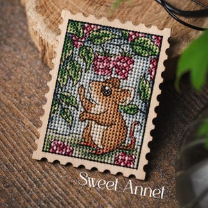 May include: A rectangular wooden plaque shaped like a postage stamp featuring a cross-stitch design of a brown mouse reaching for red raspberries on green vines. The text Sweet Annet is printed in white at the bottom of the wooden frame.