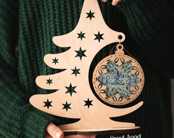 May include: A wooden Christmas tree decoration with star cutouts and a decorative ornament. The ornament features a blue and green floral design with the words "Merry Christmas" in script. The base of the tree is dark brown.