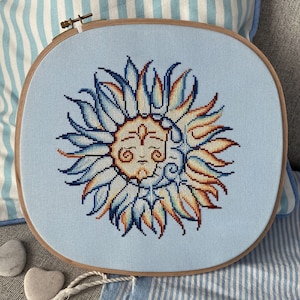 May include: A blue and orange embroidered sun with a face, framed in a wooden hoop. The hoop is on a blue and white striped fabric.