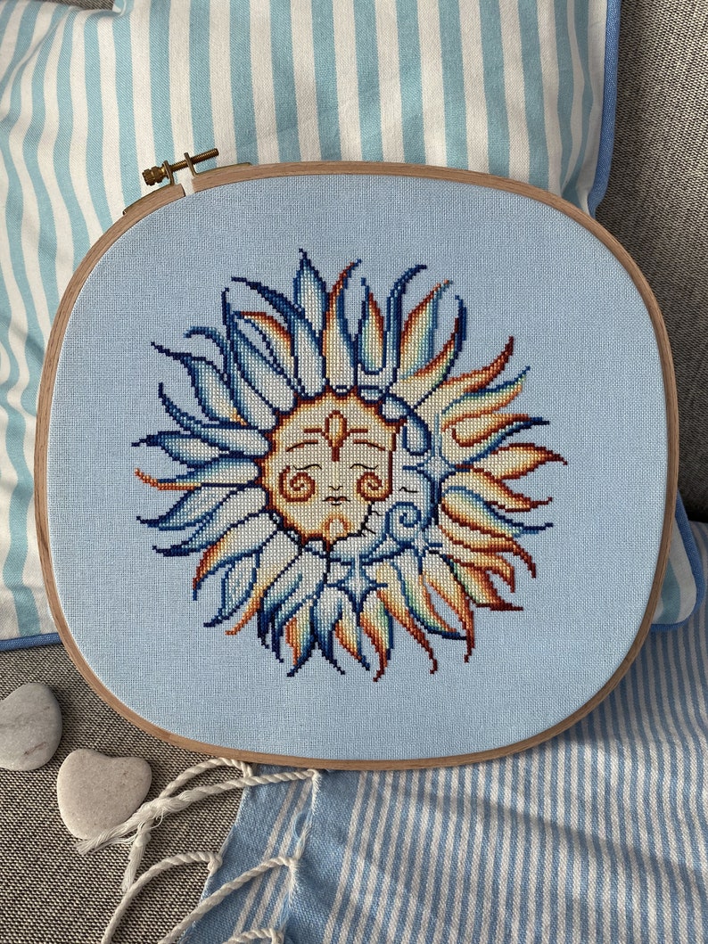 May include: A blue and orange embroidered sun with a face, framed in a wooden hoop. The hoop is on a blue and white striped fabric.