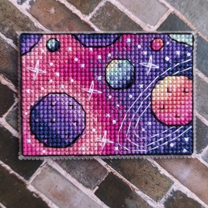 May include: A cross-stitch embroidery of a galaxy with planets and stars in shades of pink, purple, and blue.