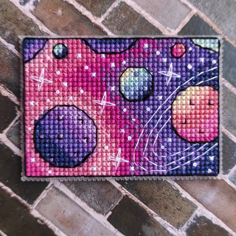 May include: A cross-stitch embroidery of a galaxy with planets and stars in shades of pink, purple, and blue.