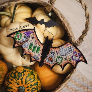 以下が含まれることがあります： 黒いコウモリ型のハロウィーンの装飾品。クロスステッチのディテールで、お化けやその他のお祝いの画像が描かれています。カボチャでいっぱいの編みかごの中にあります。「Sweet Annet」の文字が見えます。カボチャは様々なサイズと色です。