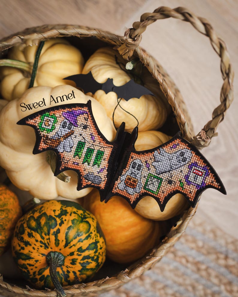 以下が含まれることがあります： 黒いコウモリ型のハロウィーンの装飾品。クロスステッチのディテールで、お化けやその他のお祝いの画像が描かれています。カボチャでいっぱいの編みかごの中にあります。「Sweet Annet」の文字が見えます。カボチャは様々なサイズと色です。