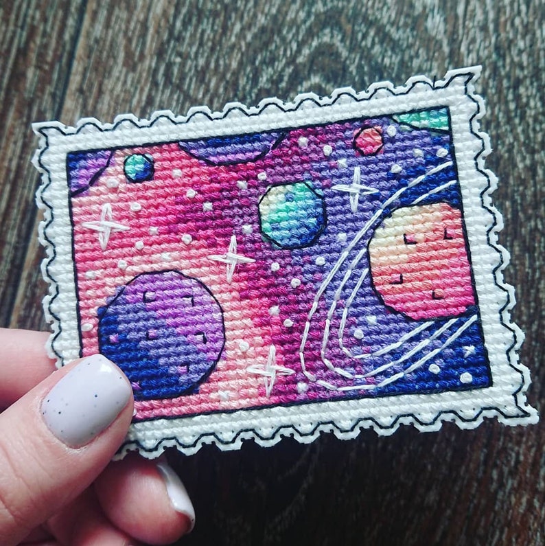 May include: A cross-stitch embroidery of a galaxy with planets, stars, and swirling nebulae in shades of pink, purple, and blue. The design is framed with a white border and a black scalloped edge.