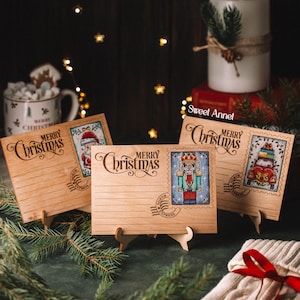 Op de afbeelding: Houten kerstpostkaarten met lasergeëtste "Merry Christmas" tekst en postzegelontwerpen. De postzegels tonen een kerstman, een notenkraker en een auto met cadeaus. De kaarten worden gepresenteerd op kleine houten standaards, met een kaars en groen in de achtergrond.