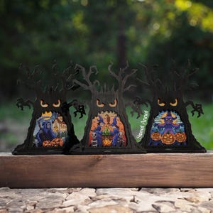 Könnte beinhalten: Drei schwarze Halloween-Baumdekorationen mit ausgeschnittenen Designs. Jeder Baum zeigt eine gruselige Szene mit Kürbissen und Figuren. Die Bäume werden auf einer Holzoberfläche vor einem verschwommenen grünen Hintergrund ausgestellt.