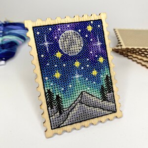 May include: A wooden stamp with a cross-stitch design of a night sky with a full moon, stars, and mountains. The stamp is surrounded by a decorative border.