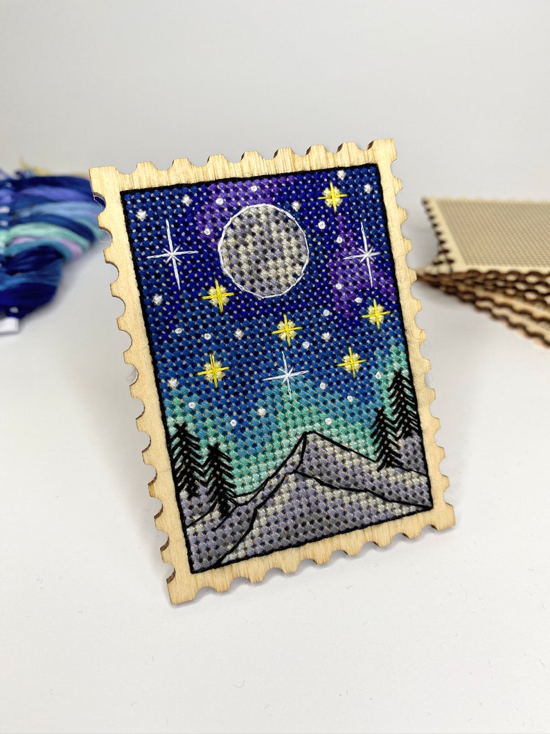 May include: A wooden stamp with a cross-stitch design of a night sky with a full moon, stars, and mountains. The stamp is surrounded by a decorative border.