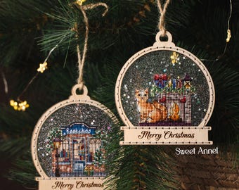 May include: Two wooden Christmas ornaments with cross-stitch designs. One features a bookstore, the other a cat with gifts. Both have "Merry Christmas" on the base and hang from twine. The ornaments are set against a backdrop of a Christmas tree.