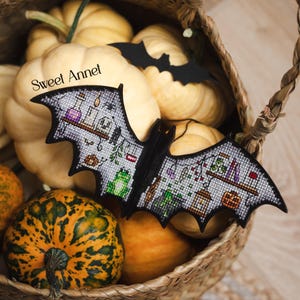 以下が含まれることがあります： ポーションボトル、カエル、その他のハロウィーンをテーマにした要素が特徴の、黒いコウモリ型の装飾品。 コウモリは、オレンジ色と黄色のカボチャなど、さまざまな形と色のカボチャが入ったバスケットの中に置かれています。 テキスト「Sweet Annet」が表示されています。