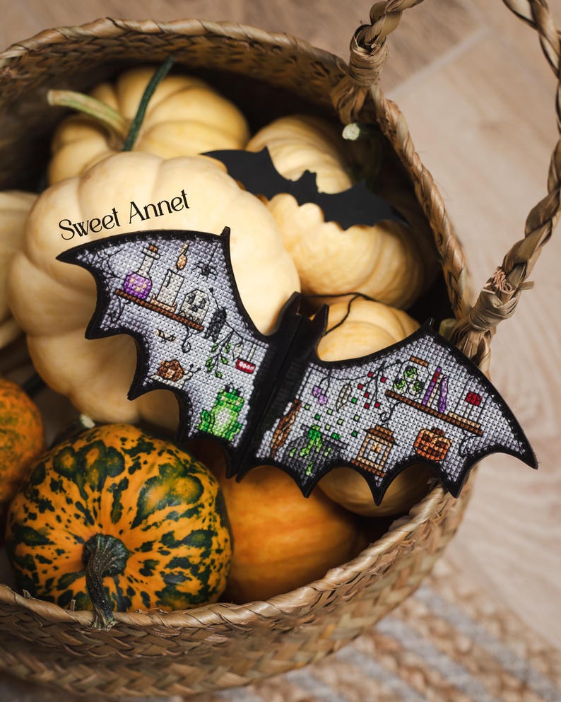以下が含まれることがあります： ポーションボトル、カエル、その他のハロウィーンをテーマにした要素が特徴の、黒いコウモリ型の装飾品。 コウモリは、オレンジ色と黄色のカボチャなど、さまざまな形と色のカボチャが入ったバスケットの中に置かれています。 テキスト「Sweet Annet」が表示されています。