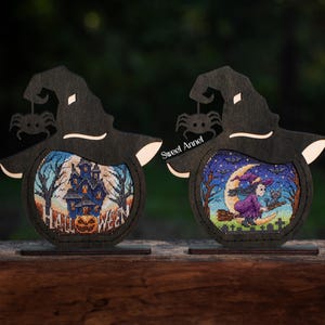 Puede incluir: Dos decoraciones de Halloween de madera con forma de sombreros de bruja, cada una con un recorte circular que muestra una escena de punto de cruz. Uno muestra una casa embrujada, el otro una bruja volando en una escoba. La palabra "HALLOWEEN" es visible.