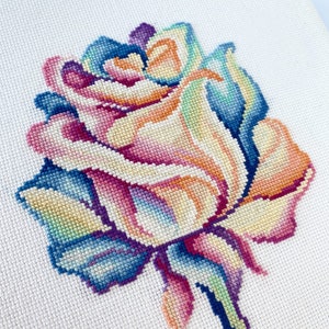 Pode incluir: Um bordado de ponto cruz colorido de uma única rosa com pétalos em tons de rosa, azul, amarelo e verde.