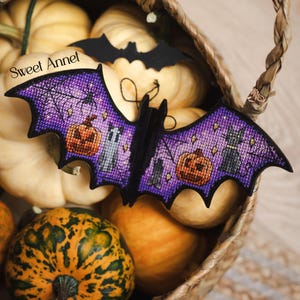 以下が含まれることがあります： 紫色の翼を持つハロウィーンのコウモリの装飾品。カボチャ、黒猫、クモの巣のクロスステッチデザインが施されています。 コウモリはカボチャでいっぱいの籐かごの中に置かれ、背景には黒いコウモリの切り抜きがあります。