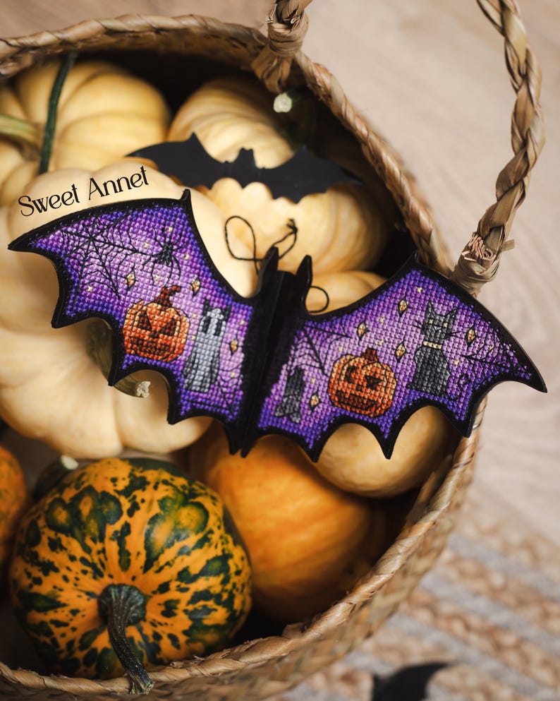 以下が含まれることがあります： 紫色の翼を持つハロウィーンのコウモリの装飾品。カボチャ、黒猫、クモの巣のクロスステッチデザインが施されています。 コウモリはカボチャでいっぱいの籐かごの中に置かれ、背景には黒いコウモリの切り抜きがあります。