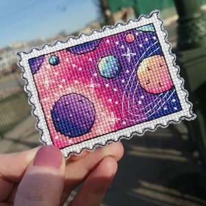 May include: A colorful cross-stitch embroidery of a space scene with planets, stars, and a galaxy background. The embroidery is framed with a white border and a decorative edge.