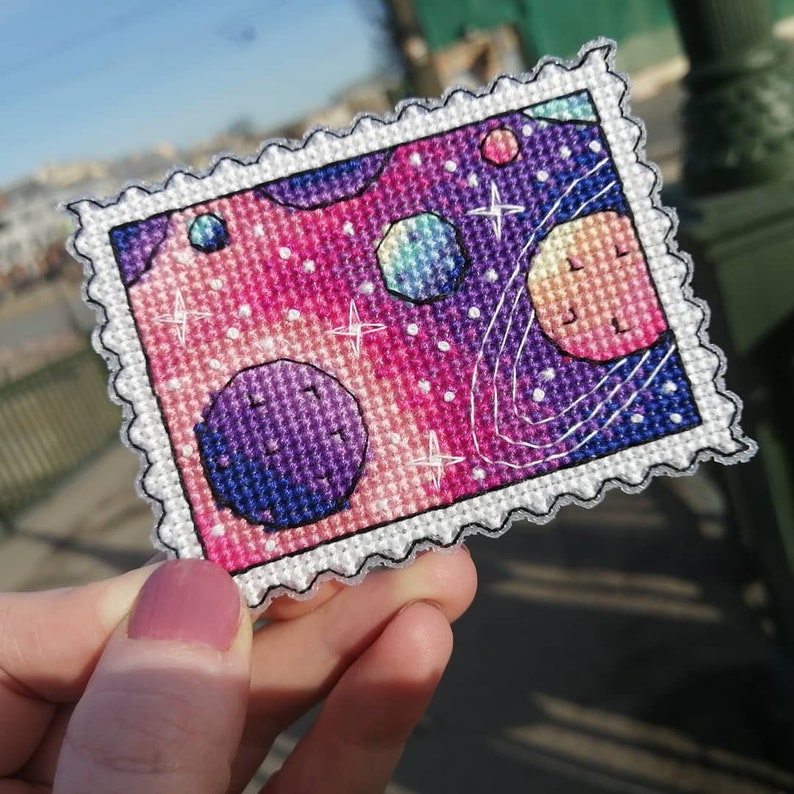May include: A colorful cross-stitch embroidery of a space scene with planets, stars, and a galaxy background. The embroidery is framed with a white border and a decorative edge.