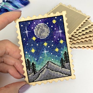 May include: A wooden craft kit featuring a cross-stitch design of a night sky with a full moon, stars, and mountains. The kit includes a pre-cut piece of wood with a scalloped edge and a piece of Aida cloth.