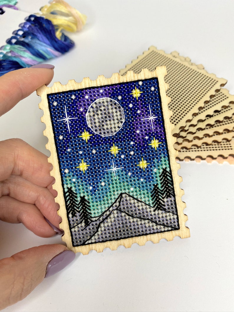 May include: A wooden craft kit featuring a cross-stitch design of a night sky with a full moon, stars, and mountains. The kit includes a pre-cut piece of wood with a scalloped edge and a piece of Aida cloth.