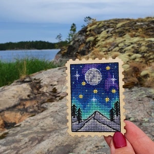 May include: A wooden stamp with a cross-stitch design of a mountain scene under a full moon and stars. The sky is a gradient of blue, purple, and green.