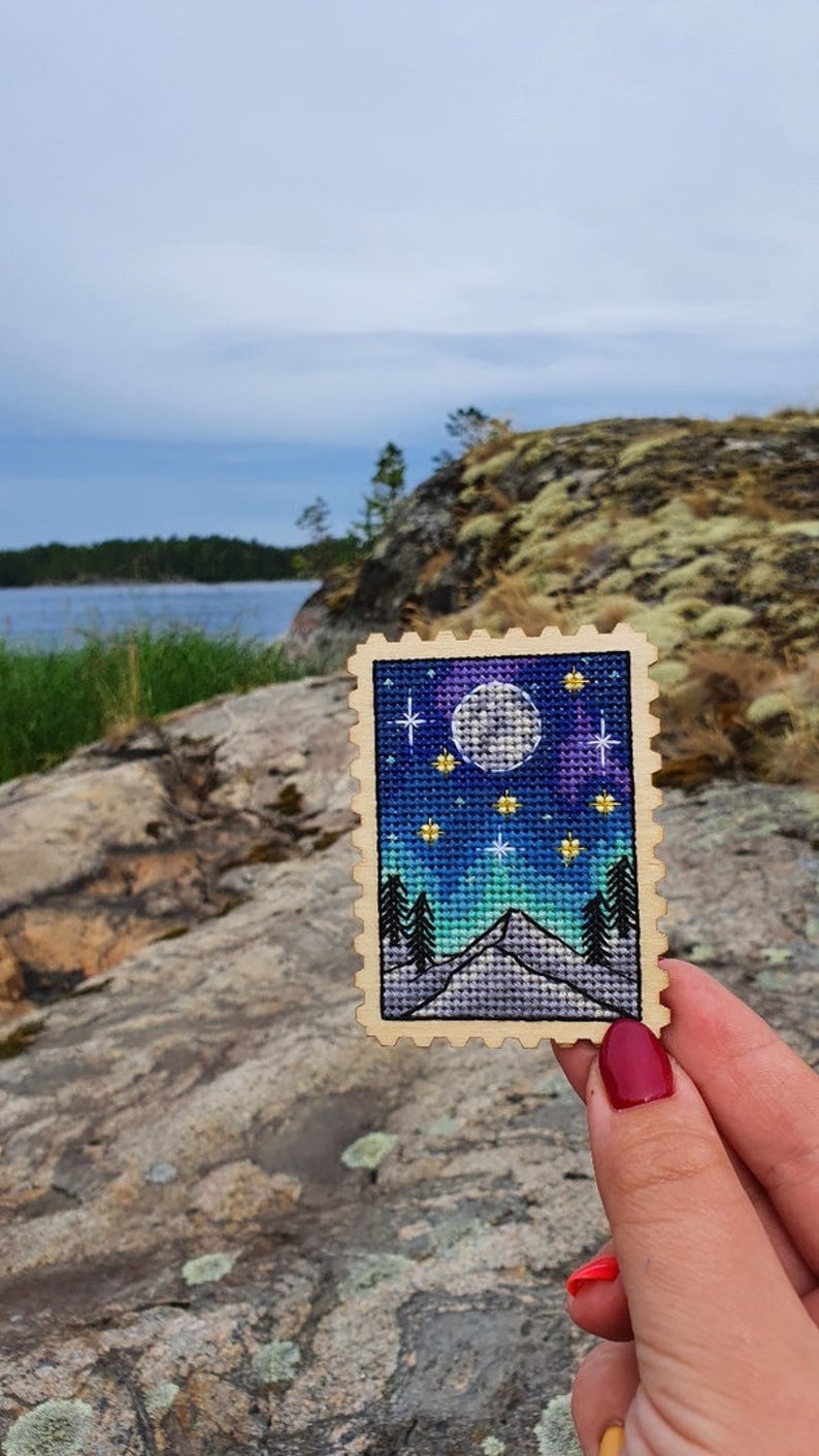 May include: A wooden stamp with a cross-stitch design of a mountain scene under a full moon and stars. The sky is a gradient of blue, purple, and green.