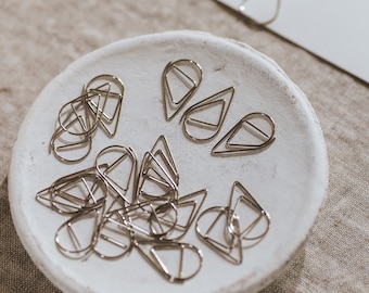 May include: A white ceramic dish filled with silver paper clips shaped like teardrops. The clips are arranged in a random pattern.