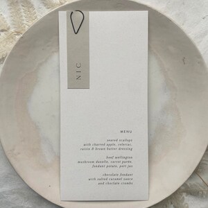 May include: A white card with a black clip and the word "NIC" printed on it. The card has a menu printed on it with the following items: seared scallops with charred apple, celeriac, raisin & brown butter dressing, beef wellington, mushroom duxelle, carrot pur&eacute;e, fondant potato, port jus, chocolate fondant with salted caramel sauce and chocolate crumbs.