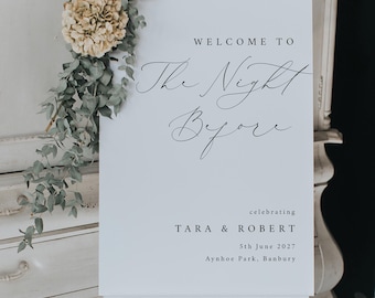 May include: A white welcome sign on an easel, with the words "WELCOME TO The Night Before" in elegant script. The sign also includes the names "TARA & ROBERT", the date "5th June 2027", and the location "Aynhoe Park, Banbury". Decorative flowers and greenery are arranged above the sign.
