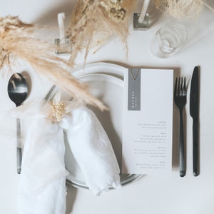 May include: A white plate with a white napkin tied with a small bunch of dried flowers. A menu card with the text "Rachel" and a description of the meal is on the plate. A black fork and knife are on the right side of the plate.