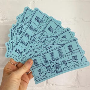 May include: A collection of light blue stickers featuring a detailed illustration of a building with windows, a door, and surrounding trees. The stickers have a dark blue outline and are held in a hand.