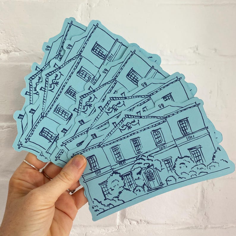 May include: A collection of light blue stickers featuring a detailed illustration of a building with windows, a door, and surrounding trees. The stickers have a dark blue outline and are held in a hand.