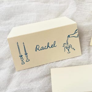 May include: A beige place card with blue ink. The card has a drawing of two candlesticks, the name "Rachel" written in cursive, and a drawing of a bottle pouring wine into a glass.