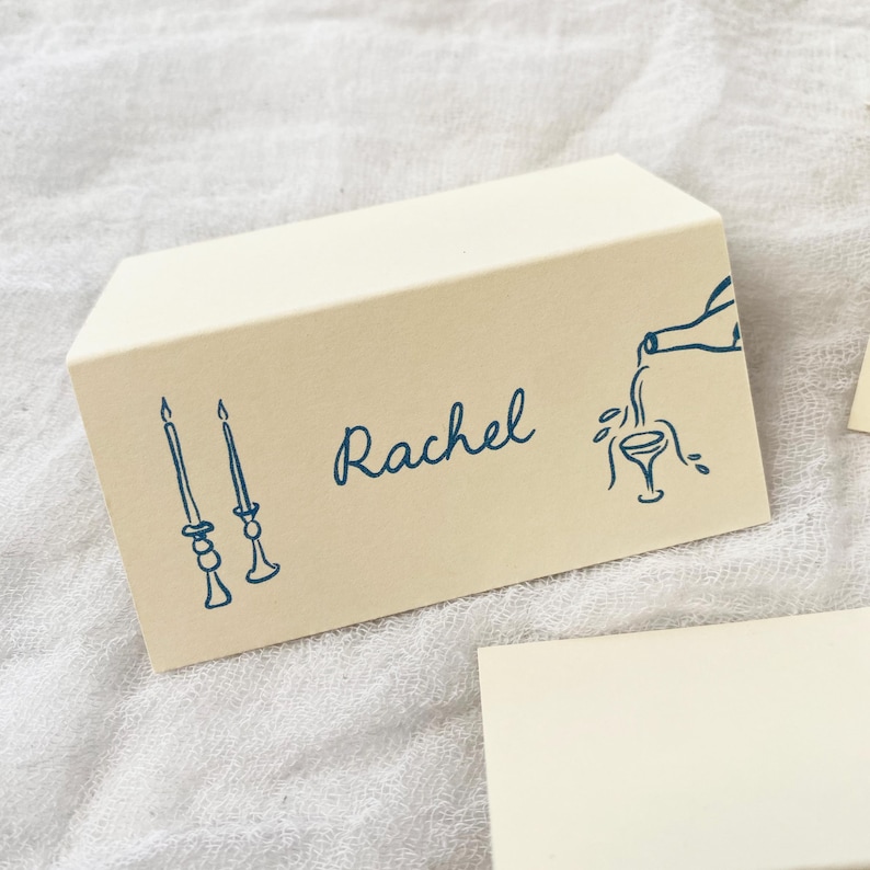 May include: A beige place card with blue ink. The card has a drawing of two candlesticks, the name "Rachel" written in cursive, and a drawing of a bottle pouring wine into a glass.