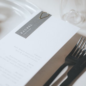 May include: A white menu with the text "RACHEL vegetarian" printed on a gray rectangle. A gold heart-shaped paper clip is attached to the top of the rectangle. A black fork and knife are placed on the table next to the menu.