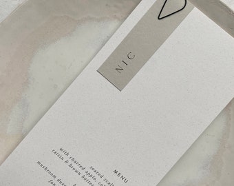 May include: A white menu card with the word "MENU" and the name "NIC" printed on it. The menu lists a dish of seared scallops with charred apple, celeriac, raisin and brown butter dressing, and a beef wellington with mushroom duxelle, carrot puree, fondant potato and chocolate.