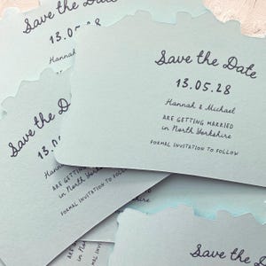 May include: A stack of light blue "Save the Date" cards with a unique wavy edge. The cards feature handwritten text, including the date "13.05.28", names, and location details for a wedding in North Yorkshire.