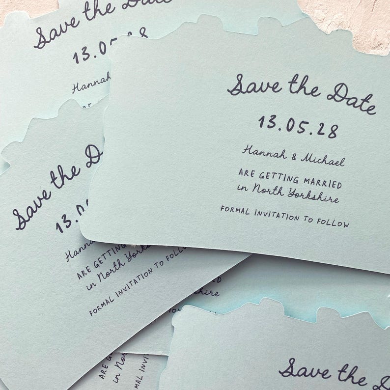 May include: A stack of light blue "Save the Date" cards with a unique wavy edge. The cards feature handwritten text, including the date "13.05.28", names, and location details for a wedding in North Yorkshire.
