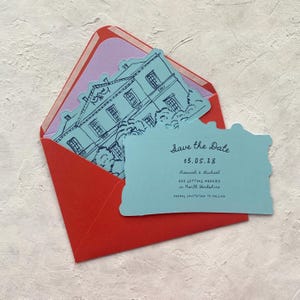 May include: A red envelope with a light purple interior holds a blue card with a house illustration and the text "Save the Date". The card also includes the date 13.05.28 and the names Hannah & Michael.