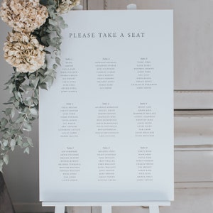 May include: A white sign with blue text lists tables and guest names for a wedding reception. The sign is on a white easel and has the words "Please Take a Seat" at the top.